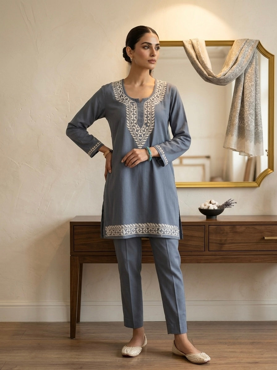 A woman models a two-piece traditional outfit consisting of a dusty blue-gray kurta with intricate white embroidery around the neckline and hem, paired with matching slim-fit trousers. She is standing in a styled room with a wooden console table and a mirror in the background.