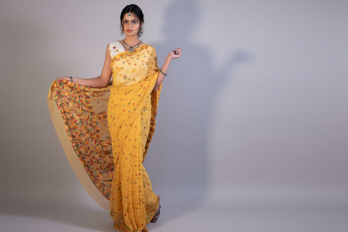 A woman posing in a yellow colour Georgette Saree with chikankari Keel Jaal Embroidery and Resham Work, with a red blouse piece and traditional Indian jewellery.