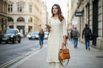 A woman standing on a sidewalk with a suitcase, wearing a cream colour traditional Lucknowi chikankari kurti with floral patterns and palazzo pants, holding a mobile phone and a bag.
