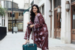 A woman standing on a sidewalk with a suitcase, wearing a traditional Lucknowi chikankari kurti with floral patterns and palazzo pants, holding a mobile phone and a bag.