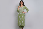 A woman standing on a sidewalk with wearing a green pink colour traditional Lucknowi chikankari kurti with floral patterns and palazzo pants, holding a mobile phone and a bag.
