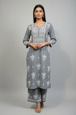 A woman standing on a sidewalk with wearing a grey colour traditional Lucknowi chikankari kurti with floral patterns and palazzo pants, holding a mobile phone and a bag.