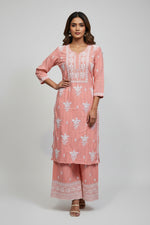 A woman standing on a sidewalk with wearing a peach pink colour traditional Lucknowi chikankari kurti with floral patterns and palazzo pants, holding a mobile phone and a bag.