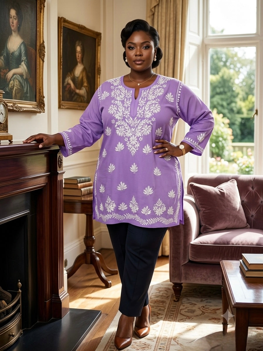 A woman stands in a classic indoor room wearing a lavender purple tunic top with white floral embroidery, paired with dark trousers and brown boots.