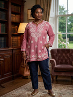 A woman stands in a furnished room wearing a dusty pink embroidered tunic top with white floral patterns, paired with navy blue pants and gold flats. She holds a brown handbag, with a bookshelf, lamp, and tufted armchair visible in the background.