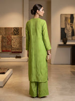 A woman stands with her back to the camera wearing a bright lime green embroidered kurta and matching wide-leg trousers in a modern, well-lit gallery space.