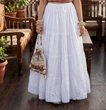 A woman wearing a long white tiered maxi skirt with floral lace detailing and a drawstring waist, standing in a rustic indoor setting with wooden furniture and holding a small embroidered bucket bag.