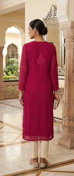 A woman wearing a magenta three-piece salwar kameez suit with cream pants and beige shoes, standing in a grand indoor courtyard with stone pillars and arches.