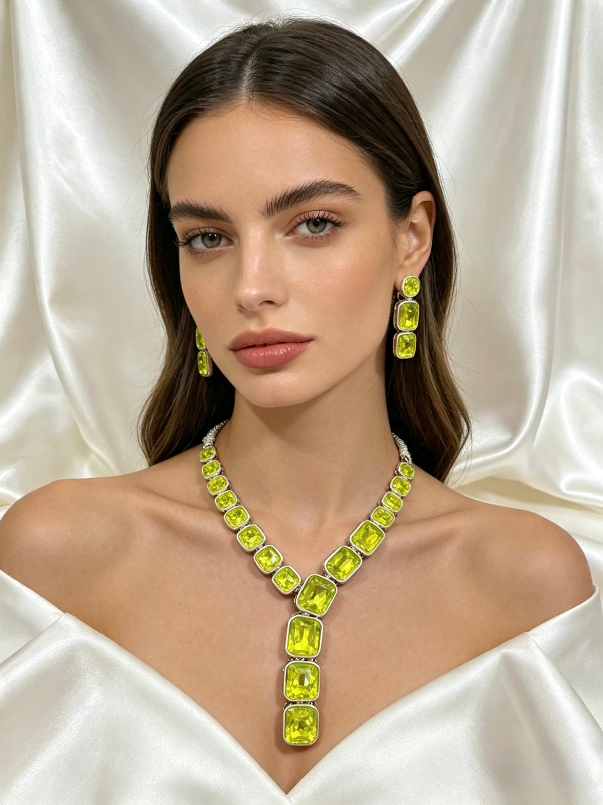 A woman wearing a neon green gemstone necklace and matching drop earrings, posed against a draped white satin fabric background.