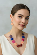 A young woman wearing the Facet Scalloped Disc Necklace, showcasing the unique pinwheel texture of the circular resin beads. The vibrant peach, ruby, and cobalt tones pop against her outfit, paired with matching peach disc drop earrings for a fresh, summer-inspired look.