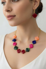 A young woman wearing the Radiant Rhombus Resin Necklace, featuring jewel-toned diamond-shaped beads in magenta, emerald, and cobalt. She is modeling the matching deep ruby-red rhombus drop earrings, which complement her vibrant, bohemian-inspired look against a soft-focus background.