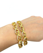 On-wrist display of two gold-toned bangles featuring an interlocking oval swirl design studded with sparkling white American Diamonds.