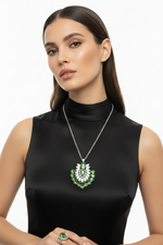 A woman wearing a silver-tone necklace with a green and white floral pendant, matching green and white drop earrings, and a green ring, styled with a black satin mock-neck top against a white background.