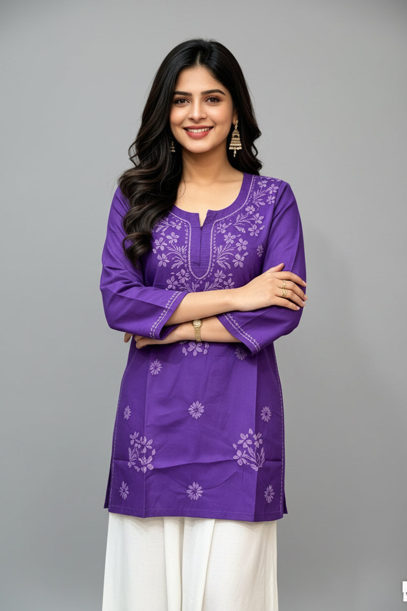 Front view of a woman wearing a royal purple cotton short kurti with white floral embroidery, a keyhole neckline, and a square hem line, paired with white trousers and standing with arms crossed.
