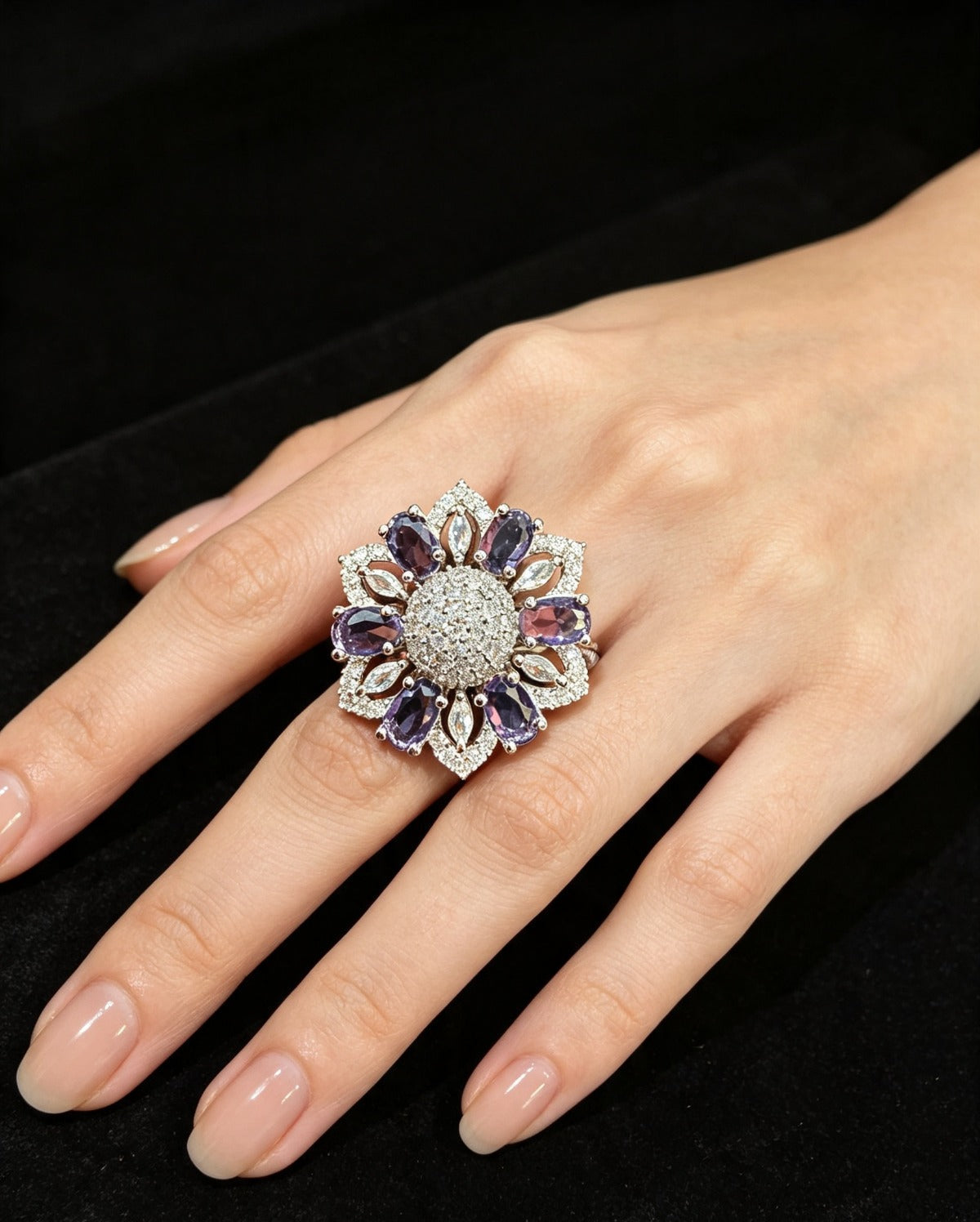 Silver-tone floral halo ring with oval purple stones, round AD center, and marquise accents — shown on hand against black velvet.