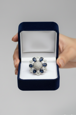 A hand holds an open dark blue velvet ring box containing a floral-shaped ring with a silver-toned center and surrounding blue stones and clear stones, against a light gray background.