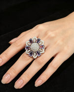 Silver-tone floral halo ring with oval purple stones, round AD center, and marquise accents — shown on hand against black velvet.