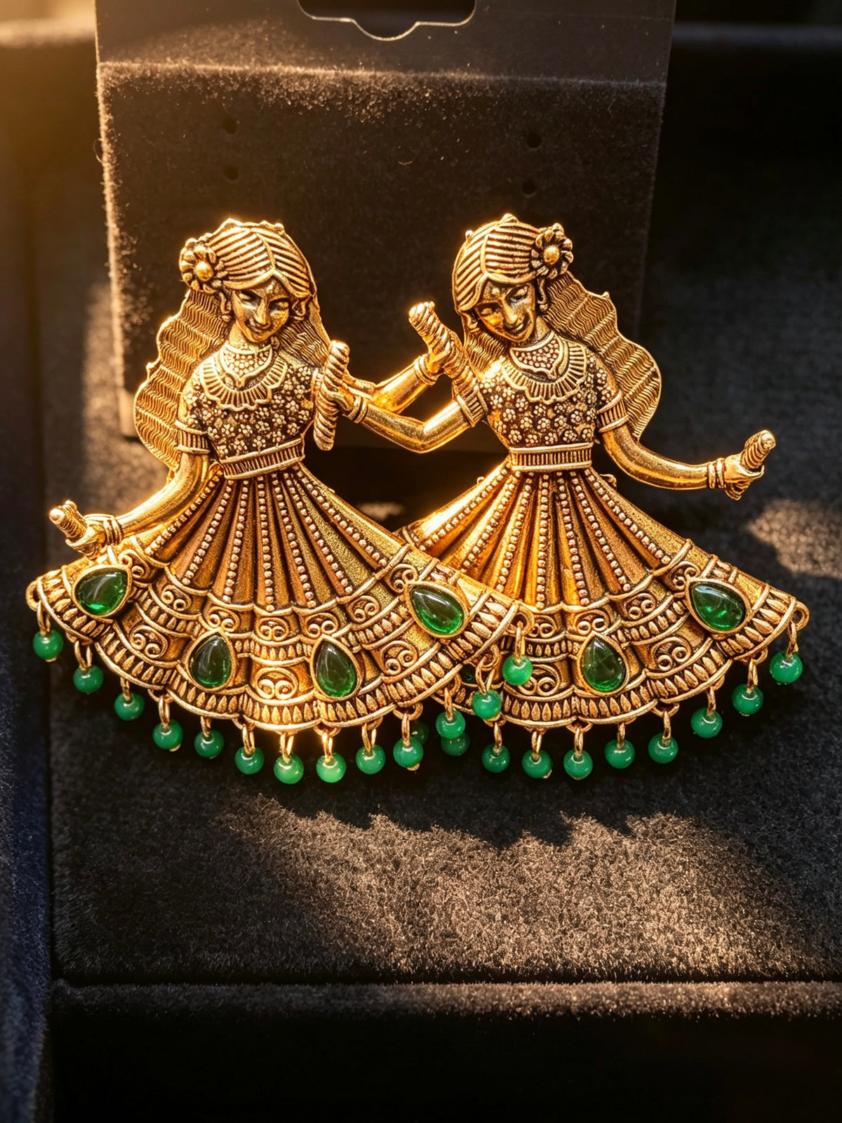 An Antique gold-toned earrings shaped like two dancing figures in navratri attire, embellished with green teardrop stones and dangling green beads, displayed on a black velvet jewellery box.