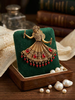 An ornate gold-toned dancing doll earrings in a decorated dress with red gemstone accents, displayed on a dark green velvet stand inside a wooden box with pearls nearby.