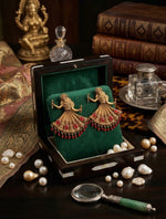 A pair of ornate gold earrings shaped like dancing figures with red gemstone drapes, displayed in an open dark wooden box lined with green velvet. The box sits on a dark wooden table surrounded by decorative props including pearls, a magnifying glass, books, and glassware.