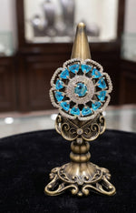 An ornate gold-toned jewellery display stand holding a decorative piece with a central turquoise-blue gemstone and surrounding clear stones, set on a black surface with a blurred jewellery store background.