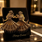 An ornate gold-toned jewellery piece depicting two dancing figures in detailed traditional attire, displayed on a black velvet stand over a polished dark marble surface.