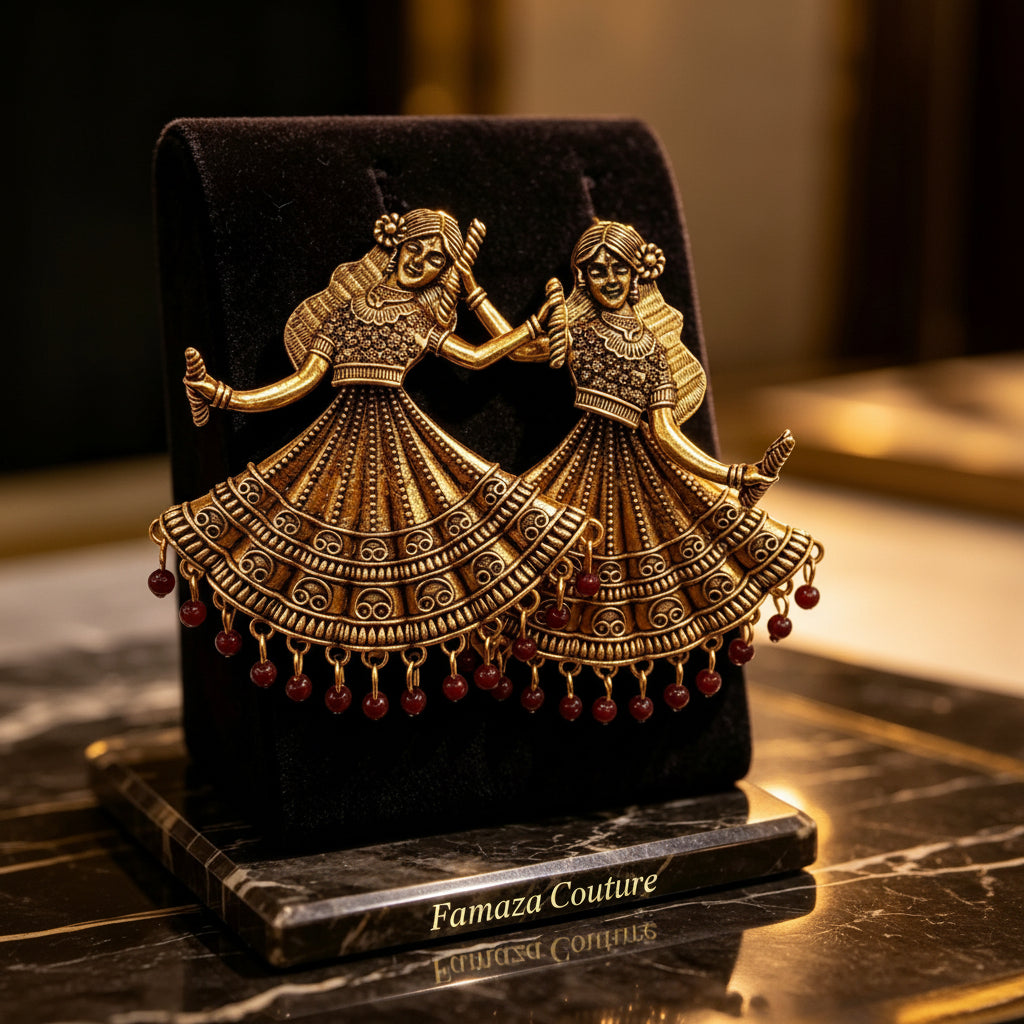 An ornate gold-toned jewelry display stand shaped like two dancing figures in traditional attire, holding hands and wearing layered embroidered dresses with dangling red bead drops, set on a black velvet cushion over a polished marble surface.