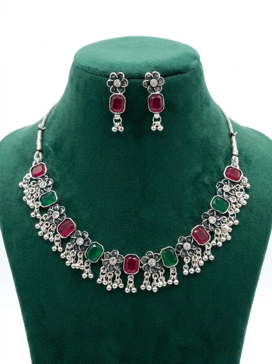 High-contrast view of an oxidized silver jewelry set with rectangular hot pink and emerald green stones and ghungroo drops on a green velvet stand.