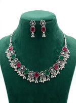 High-contrast view of an oxidized silver jewelry set with rectangular hot pink and emerald green stones and ghungroo drops on a green velvet stand.