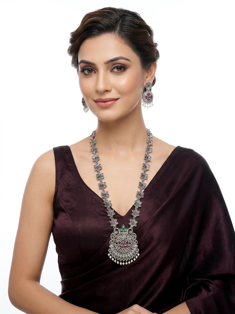 A model wearing a long-line vintage oxidized silver necklace with an intricate floral lattice design and matching drop earrings, featuring ruby and emerald-toned stone accents and white pearl droplets.