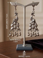 Silver-toned peacock dangle earrings displayed on a metal stand, with overlaid dimension labels for drop length, bottom width, and weight on a warm indoor background.