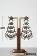 A pair of ornate silver-toned dangle earrings displayed on a wooden and metal stand, set against a dark brown draped fabric background with a blurred mirror in the distance.