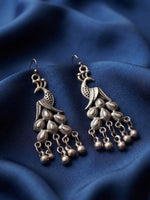 A pair of antique silver-toned peacock dangle earrings with hanging bell charms, displayed on draped dark blue fabric.