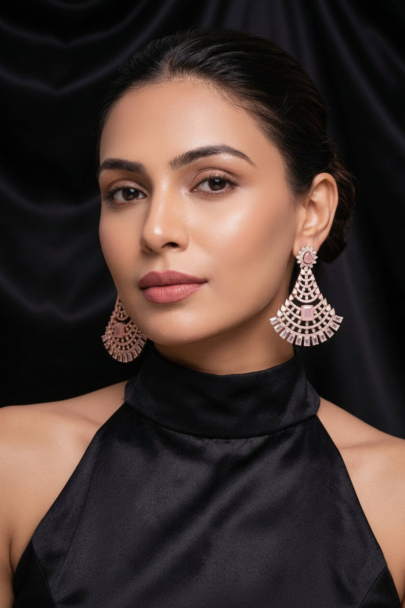 A close-up portrait of a woman wearing large ornate dangling earrings with a pink and silver-toned scalloped design, paired with a black high-neck top against a black satin background.