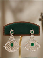 A pair of large silver-toned statement earrings with green stone centers and clear crystal fringe, displayed on a wooden jewelry stand with green velvet cushions.
