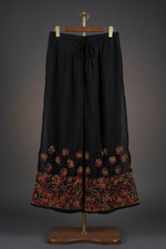 Front view of black wide-leg palazzo pants featuring vibrant multi-colored floral embroidery along the hem and an adjustable drawstring waistband.