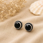 Black stud earrings with silver detailing on a beige fabric background.