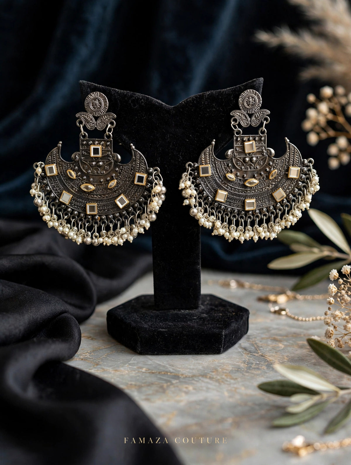 A pair of ornate silver-toned huggie earrings with intricate filigree, square gold-toned accents, and dangling pearl-like beads, displayed on a black velvet stand. The background features draped black fabric, a textured gray surface, and decorative olive branches.