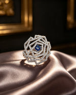 Exquisite Silver & Blue statement ring featuring a square-cut blue centre stone, intricate layered geometric diamond halo, and polished silver finish — elegantly displayed on soft champagne satin with warm golden lighting.