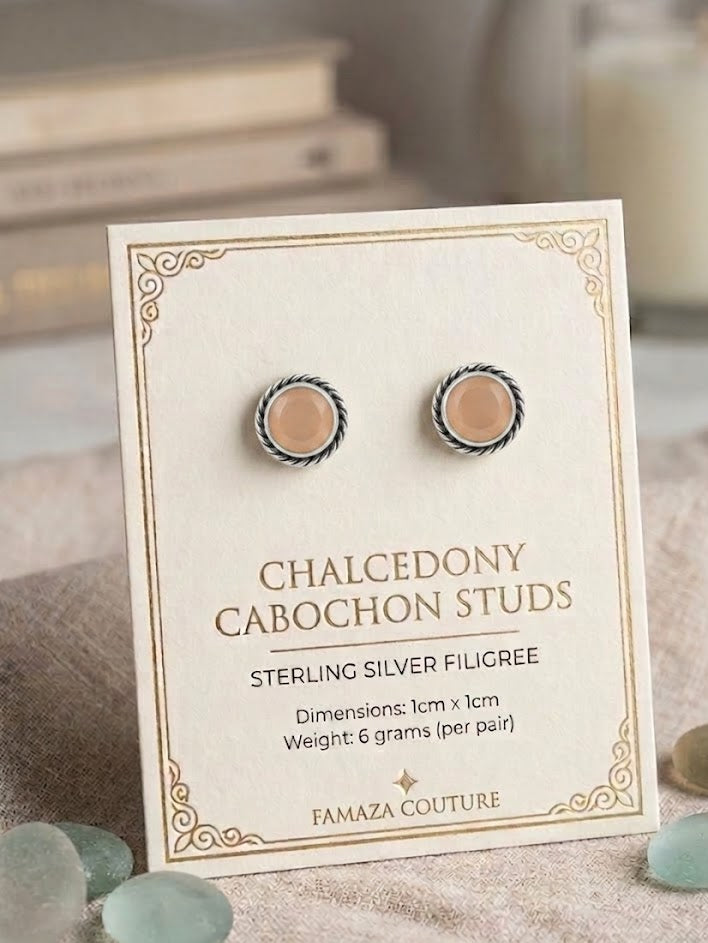 A pair of cabochon stud earrings displayed on a cream jewelry card. The round studs have a silver-toned filigree border with a warm peach-beige center.
