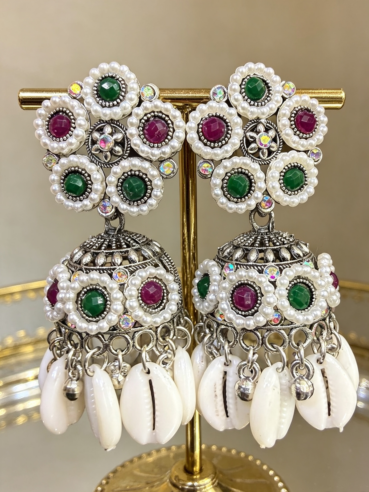 Boho-glam silver-finish jhumka earrings featuring white pearl clusters, green and maroon gemstones, and hanging white cowrie shells displayed on a gold stand.