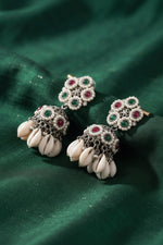 A pair of traditional Indian silver-toned dangle earrings with white cowrie shells, green and red stones, and silver filigree, displayed on a draped dark green fabric background.