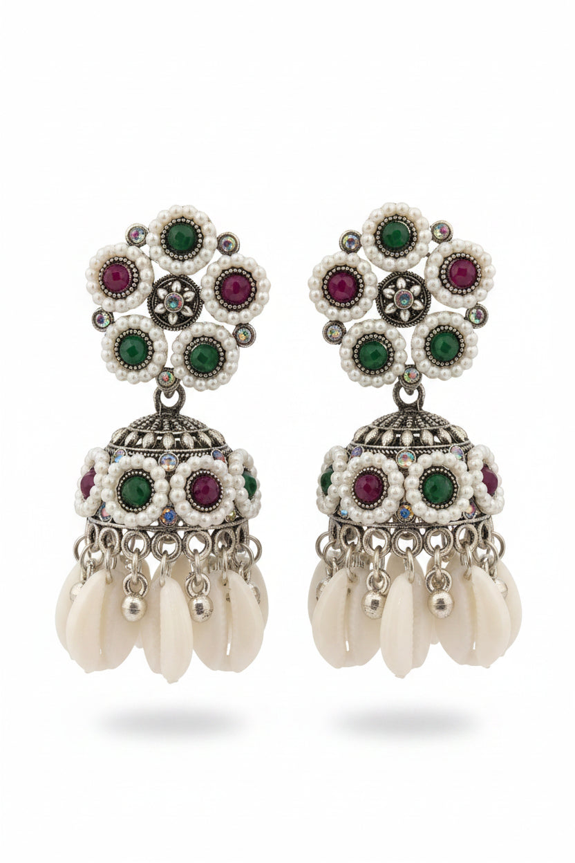 A pair of ornate silver-toned Indian jhumka earrings with white pearl-like beads, green and pink stones, and dangling white cowrie shells, held in a hand against a dark background.