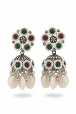 A pair of ornate silver-toned Indian jhumka earrings with white pearl-like beads, green and pink stones, and dangling white cowrie shells, held in a hand against a dark background.