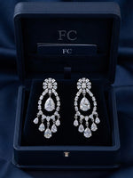 A pair of silver-toned chandelier earrings with clear teardrop gemstones, presented in an open dark navy blue jewelry box branded with 'FC'.