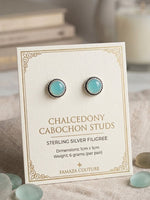 A pair of cabochon stud earrings with light blue-green stones and silver filigree settings, displayed on a cream jewelry card labeled 'Chalcedony Cabochon Studs' by Famaze Couture, styled on a linen surface with small stones and a blurred candle in the background.