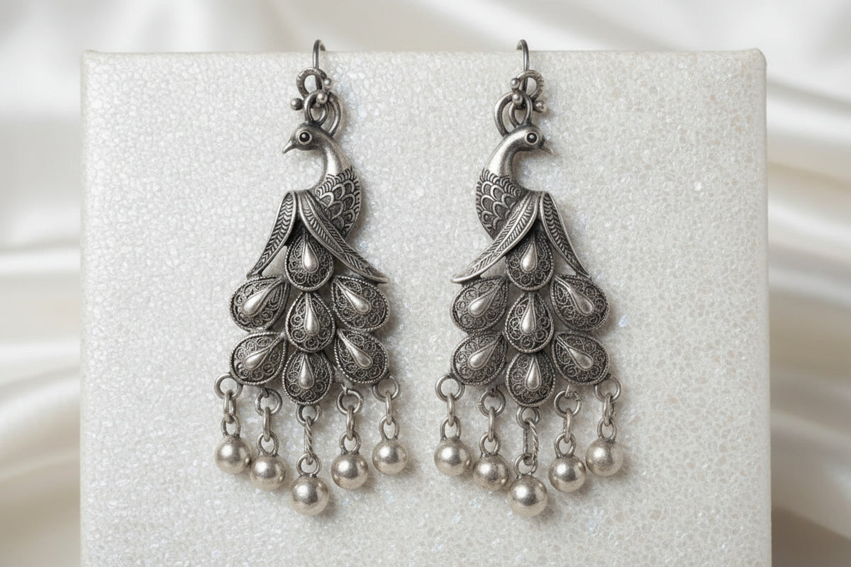 A close-up of a silver-toned traditional Indian jhumka earring worn on a person's ear. The ornate dangling earring features a peacock motif with intricate filigree detailing and hanging silver bells.