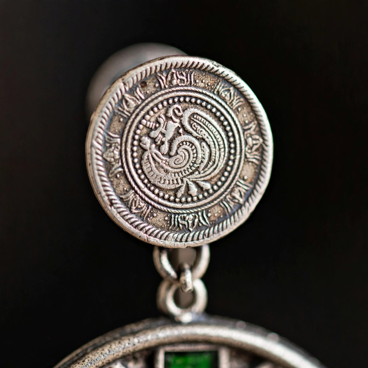 Close-up of a silver-toned coin-style earring with an engraved bird motif and a small green stone detail, shown against a dark background.