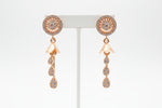 Close-up of golden brass earrings featuring a circular floral stud encrusted with crystals and a three-tier crystal teardrop dangle.