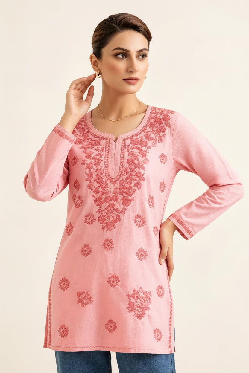 Front view of a model wearing a blush pink cotton short kurti featuring intricate pink floral embroidery, a keyhole neckline, and a square hem line, paired with matching trousers and posing with her hand touching her earring.